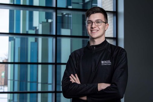 Nathan Fairweather standing and smiling, with arms crossed, in front of NEC Building windows