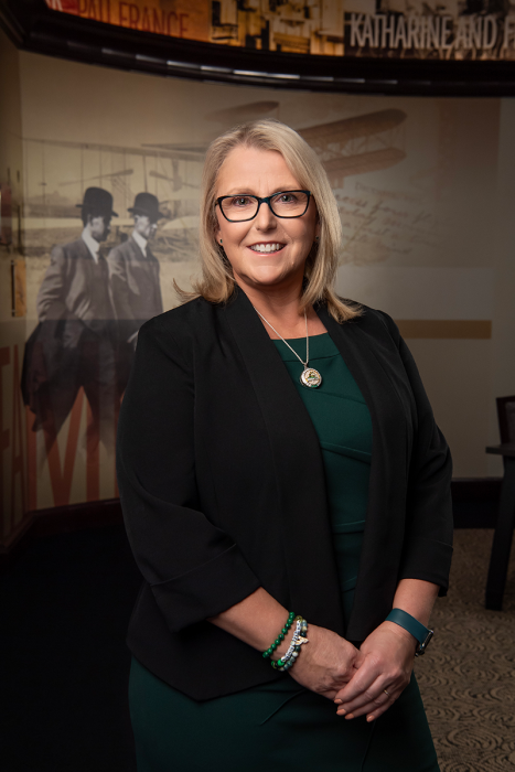 A person in business attire stands smiling at the camera. The background shows a wall with an image of Wilbur and Orville Wright and a biplane.