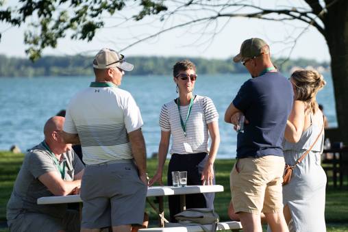 Four people stand gathered around a picnic table talking. A lake is in the background.