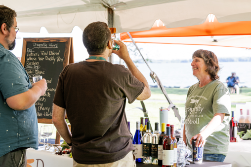 A person stands behind an outdoor booth displaying wine bottles. A man stands in front of the booth sampling a drink.