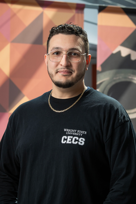 A man with glasses stands for a portrait, looking into the camera.