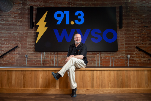 A person sitting on a wooden stage in business casual attire. The background has a large sign reading "91.3 WYSO" with a lightning bolt.