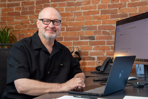 A man in a polo sitting at a desk with a computer in front of a brick wall.
