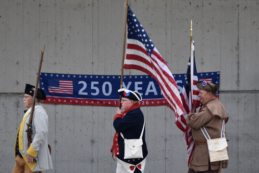 A group of people dressed in historical military uniforms carry a variety of flags, passing in front of a banner reading "250 Years."
