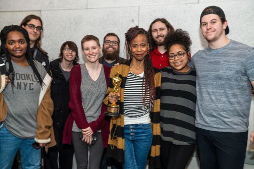 A group of people standing in front of a whiteboard, smiling at the camera. The person in the middle is holding an Oscar.