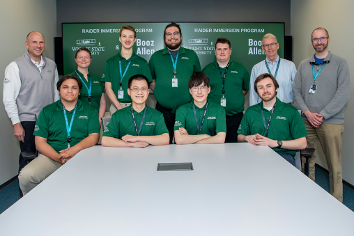 Eleven individuals sitting and standing in two rows, smiling at the camera. Most are wearing green polos. Behind them is a screen reading "Raider Immersion Program Booz Allen Hamilton."