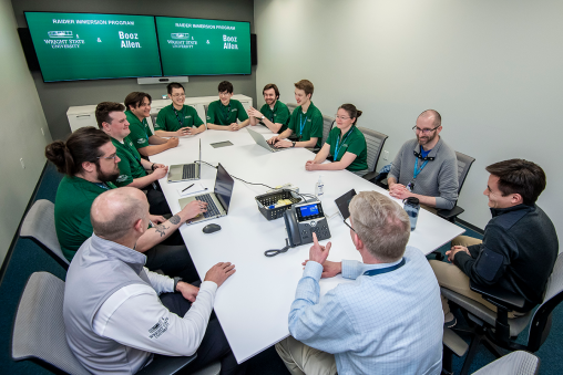 Twelve individuals sit around a table in a workplace. Most are in green polos. The digital display in the background reads "Raider Immersion Program, Wright State University x Booz Allen Hamilton."