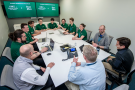 Twelve individuals sit around a table in a workplace. Most are in green polos. The digital display in the background reads "Raider Immersion Program, Wright State University x Booz Allen Hamilton."