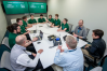 Twelve individuals sit around a table in a workplace. Most are in green polos. The digital display in the background reads "Raider Immersion Program, Wright State University x Booz Allen Hamilton."