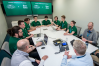 Twelve individuals sit around a table in a workplace. Most are in green polos. The digital display in the background reads "Raider Immersion Program, Wright State University x Booz Allen Hamilton."