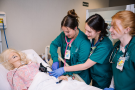 Three nursing students work on a mannequin in a hospital bed in a classroom setting.