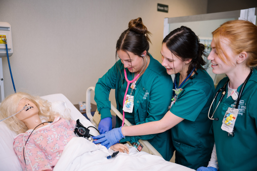 Three nursing students work on a mannequin in a hospital bed in a classroom setting.