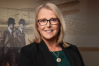 A person in business attire stands smiling at the camera. The background shows a wall with an image of Wilbur and Orville Wright and a biplane.
