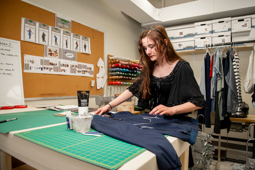 A costume designer works on a jacket. Photos are hung in the background depicting drawings of other costumes.