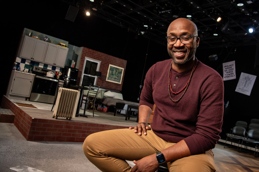 A man sits and poses for a photo. In the background is the set of a theatre production, including an apartment set.