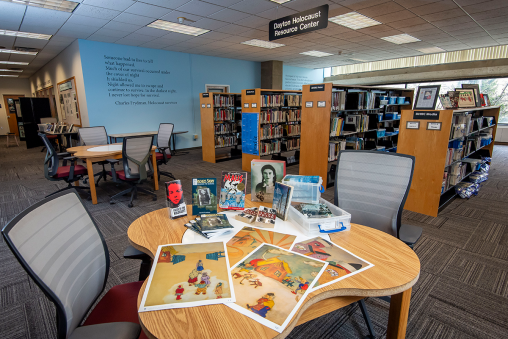 A section of a library with bookshelves lining the back. The front table has books and documents related to the Holocaust.