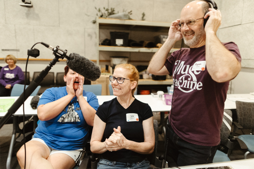 A person stands in the foreground wearing a shirt that reads "MUSE MACHINE" and headphones. Two other people sit behind them, cheering. A microphone is in view.