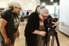 A woman stands over a film camera on a tripod, looking into it. Another person stands behind them watching.