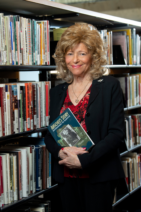 A woman standing in front of a bookshelf, holding a book titled "Anschel's Story"