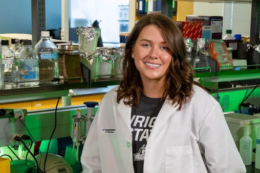 A woman in a lab coat smiles at the camera. She is in a laboratory, surrounded by equipment.