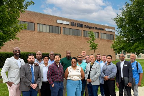 Sixteen people in business attire standing in front of a brick building. The building is labeled "Raj Soin College of Business."