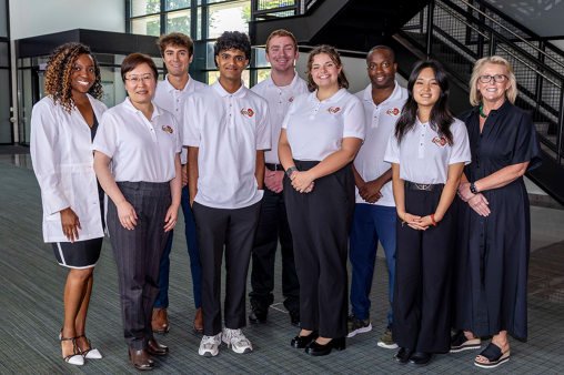 Nine individuals stand in a line, smiling for a photo. Most are in matching polos.