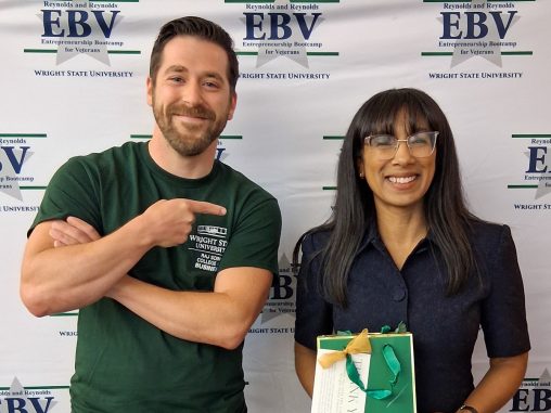 A man and woman stand together in front of a backdrop reading "EBV." The woman is holding a gift bag while the man points at her.