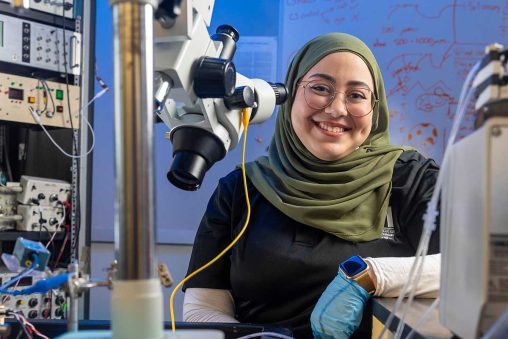 Hana Habib smiling behind the microscope in the Neuro Engineering, Rehabilitation and Degeneration lab.