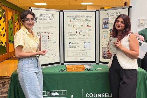 Two woman stand at a booth, covered with a tablecloth and holding up a poster board with Counseling and Wellness information on it.