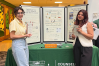 Two woman stand at a booth, covered with a tablecloth and holding up a poster board with Counseling and Wellness information on it.