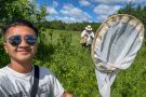 A man stands in a field full of plants, holding up a fly net. A man in the background is examining a finding.