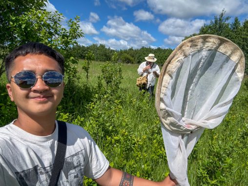 A man stands in a field full of plants, holding up a fly net. A man in the background is examining a finding.