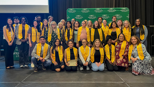 A group of students stand on a stage wearing gold graduation stoles. The backdrop behind them reads "WRIGHT STATE UNIVERSITY."