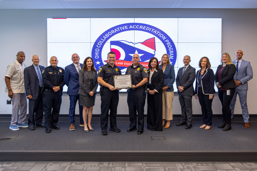 Several individuals in business attire and police officer uniforms standing together on a stage. The officers in the center are holding an award.