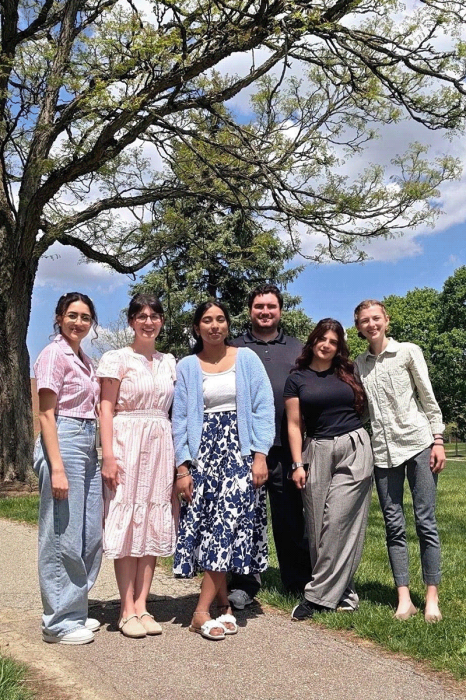 Six students stand together for a photo in an outdoor area, with trees behind them.