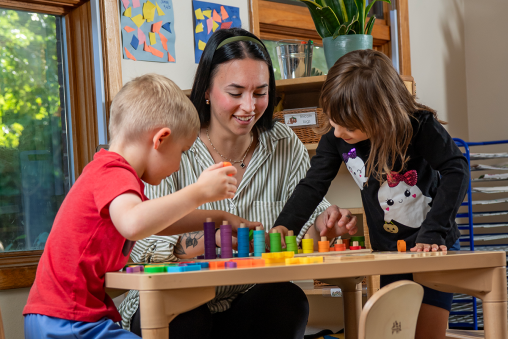 A teacher sits at a table, playing with a colorful toy with two young students.