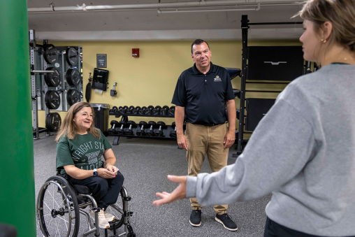 A woman in a wheelchair engages in conversation with a man and student inside a university recreation center.