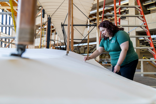 A woman in a Wright State shirt is dusting a life sized model of the Wright Flyer.