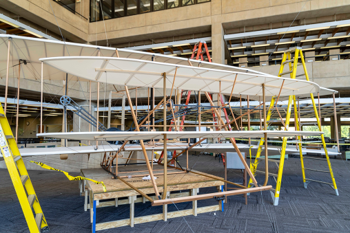 A life size model of the Wright Flyer in a library, lowered to the ground and surrounded by cleaning equipment.