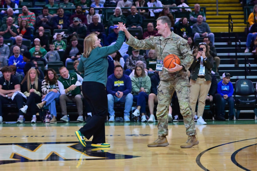 A woman high-fives a man in a military uniform. The man is holding a basketball and the two individuals are standing on a basketball court with a crowd behind them.