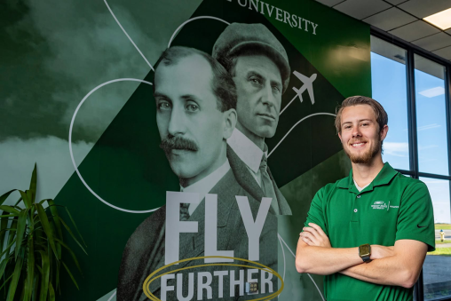 A man stands with his arms crossed by windows at an airport. The wall behind him has a graphic of the Wright brothers and says "FLY FURTHER."