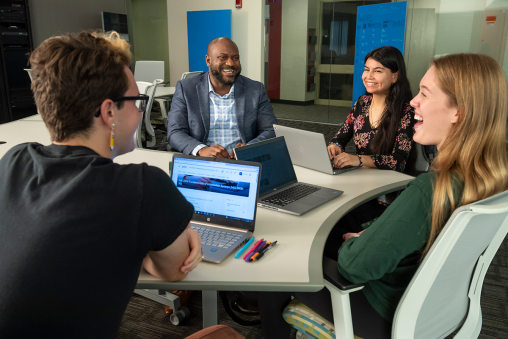 A professor and three students sit around a table together, laughing with their computers open.