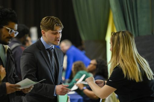A student in business attire hands a resume to an person at a career fair booth.
