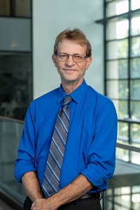 A man in business attire posting for a professional portrait in front of a wall of glass windows.