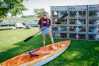 A woman stands on a paddleboard that is laying on the grass. She is wearing a life vest and holding a paddle. Behind her is a kayak rental booth.