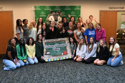 A group of high school students stand together with a sign saying "Future Teachers Day." Wright State's wolf mascot stands in the back, along with a backdrop reading "Wright State University | College of Health, Education and Human Services."