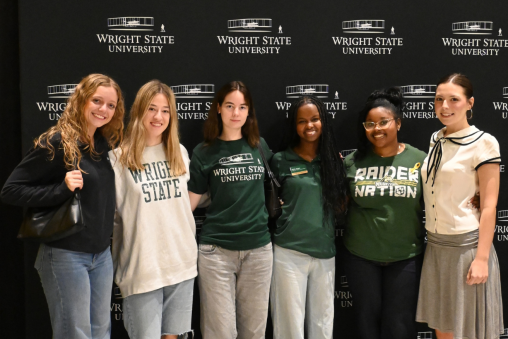 Six students stand together in front of a backdrop reading "Wright State University." Many are wearing Wright State apparel.