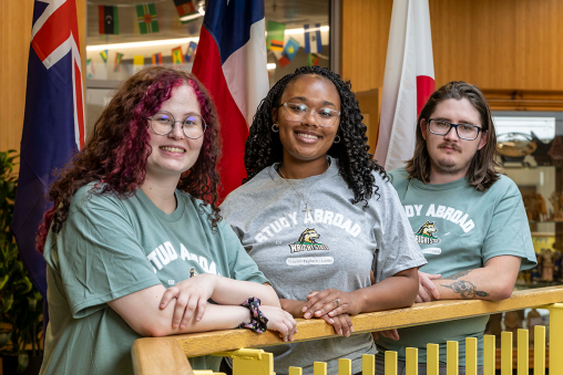 Three students, all wearing Wright State study abroad t-shirts, pose over a bannister. Different countries' flags are in the background.