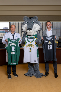 Two men and a wolf mascot stand together, each holding a Wright State Raiders basketball jersey with a Premier Health patch in the corner.